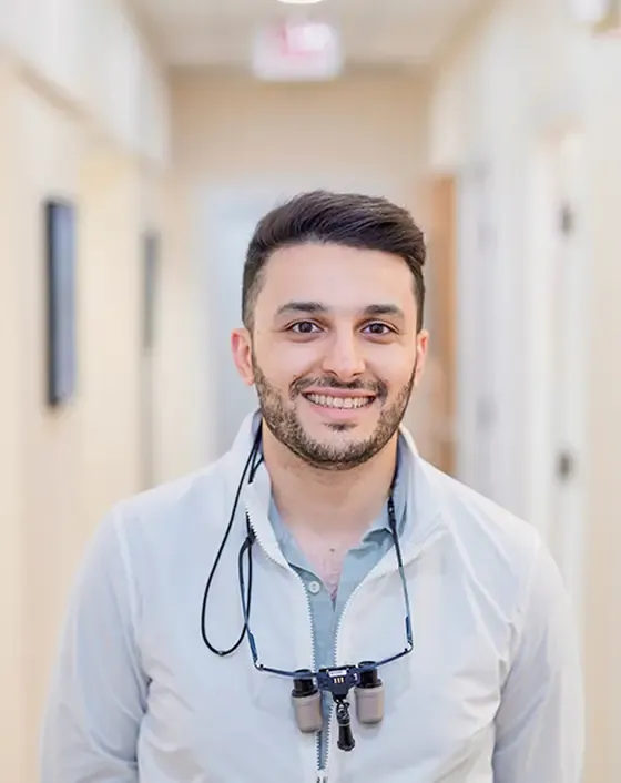 Smiling man wearing a white jacket and glasses, standing in a hallway.